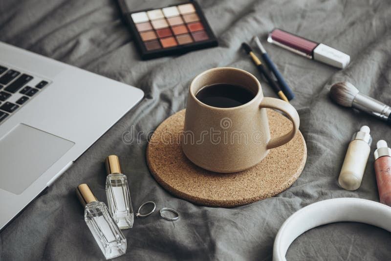 En laptop bredvid en kaffekopp och smink på en säng.